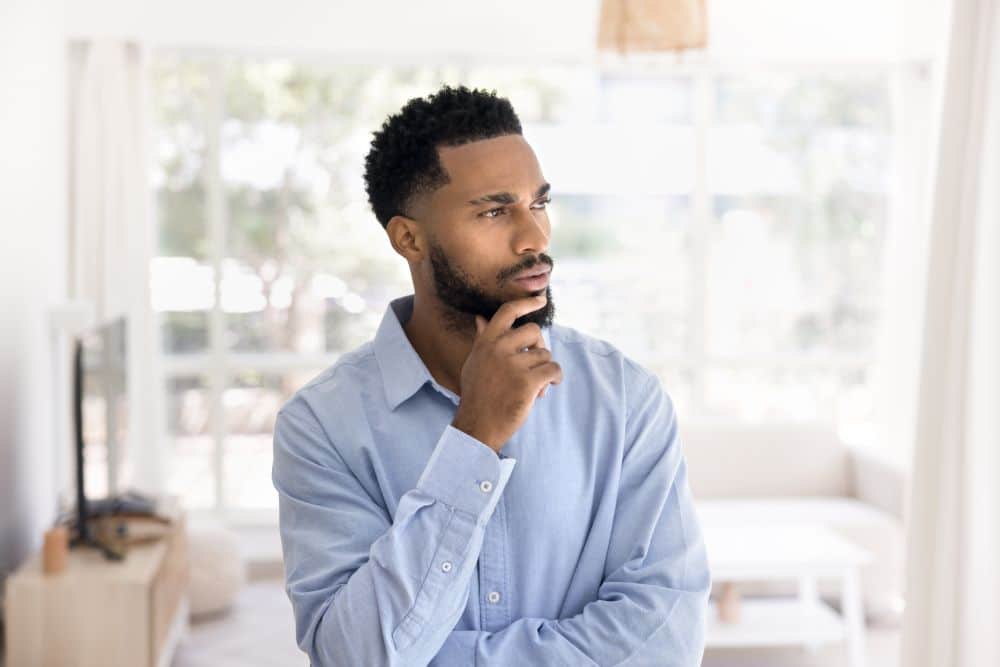 A man wearing a light blue shirt stands indoors, looking thoughtful with his hand on his chin. He is in a bright room with large windows and white furnishings in the background.