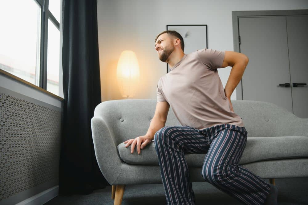 A man with severe pain in lower back on one side caused by kidney stones, trying to get up from a sofa