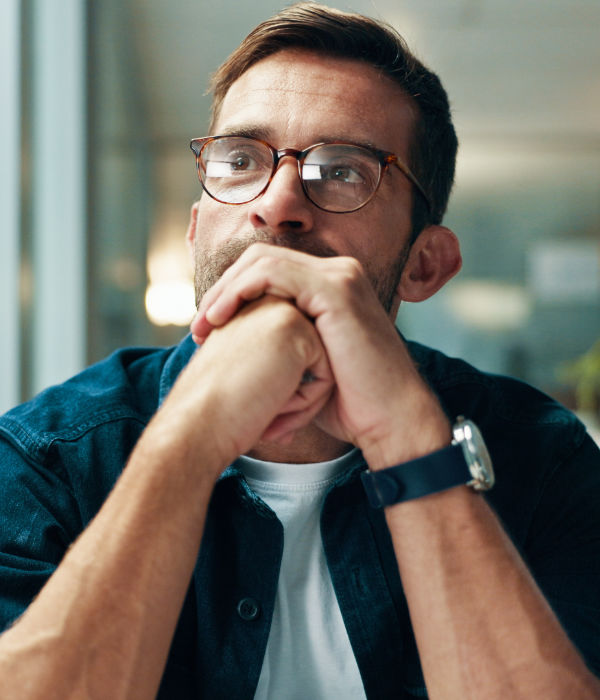 A man wearing glasses and a watch sits with his hands clasped in front of his face, looking thoughtfully into the distance.