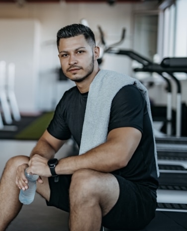 A man wearing a black t-shirt and shorts sits on gym equipment