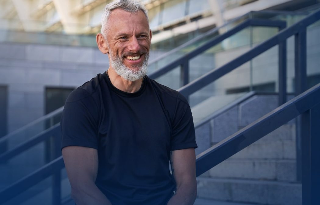 A smiling older man with short gray hair and a beard sits outdoors on steps