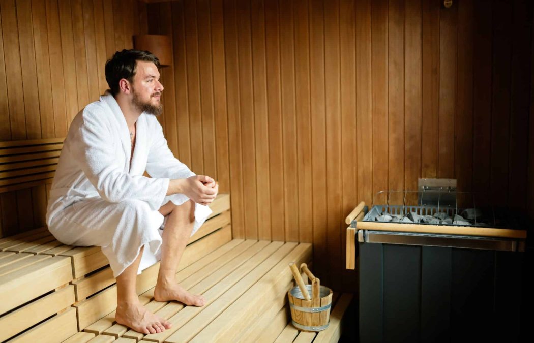 A man in a white robe sits relaxed on a wooden bench in a sauna