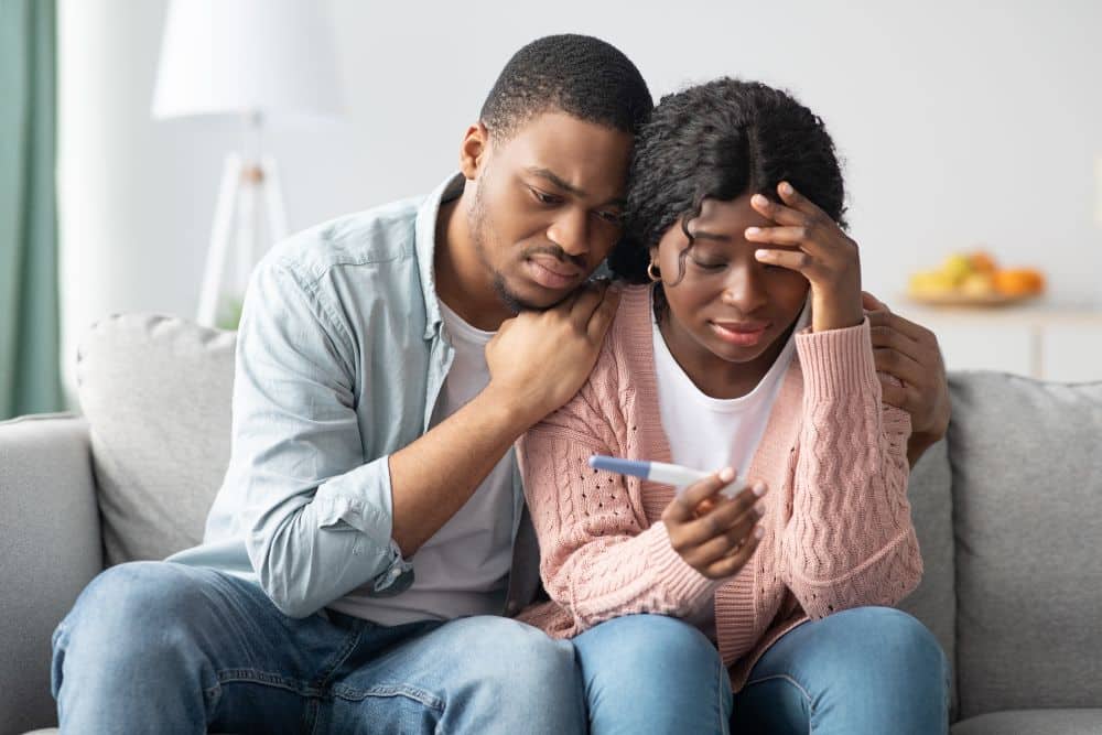 Upset Afro-American couple looking at a negative pregnancy test sitting on couch at home.