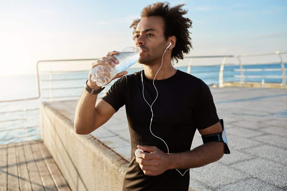 Fit afro-american male athlete drinking water out of plastic bottle after jogging on the pear.
