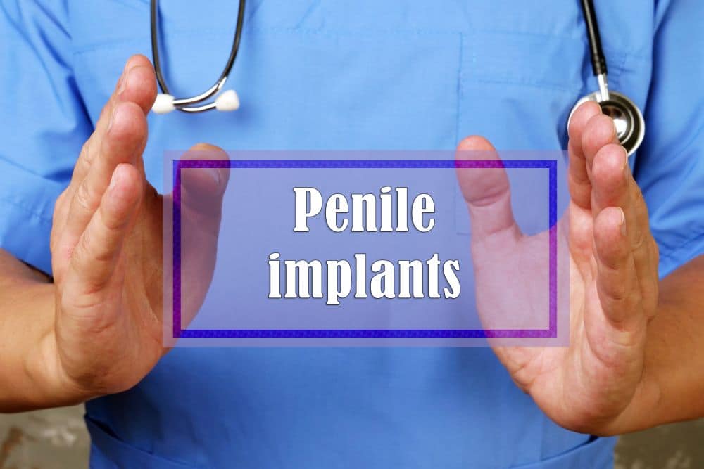 A doctor in blue scrubs and a stethoscope holds their hands apart with the words Penile implants displayed in bold white text over a blue transparent box.