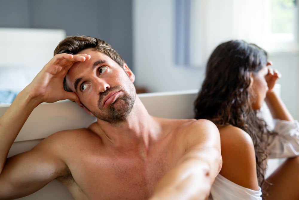 Upset and disappointed couple experiencing intimacy problems cause by man's erectile dysfunction sitting on the floor by the bed