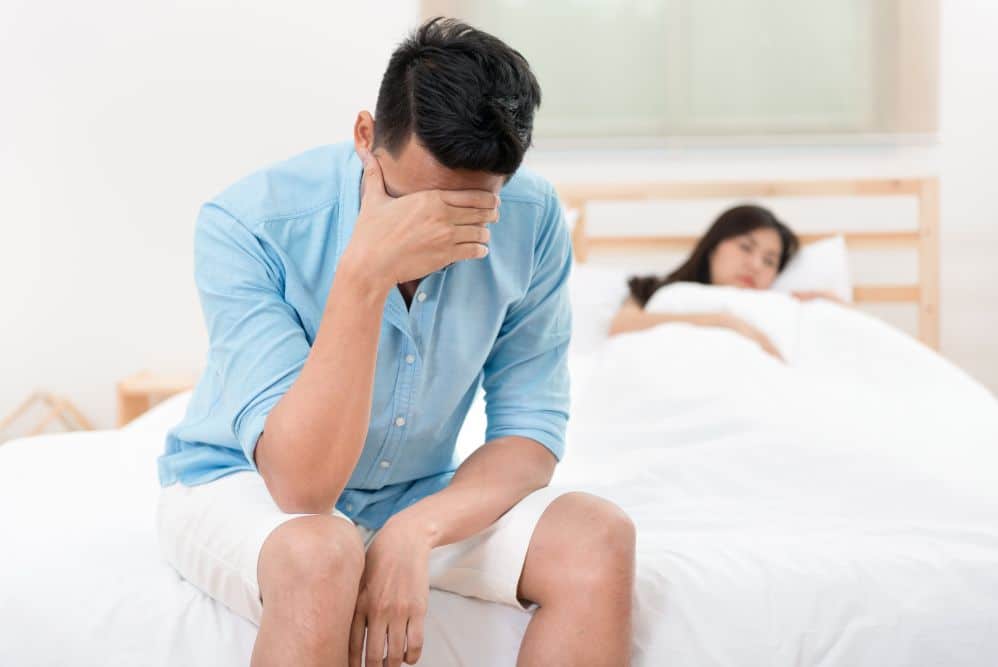 Disappointed man suffering from TRT infertility sitting on the edge of the bed next to a sleeping woman.
