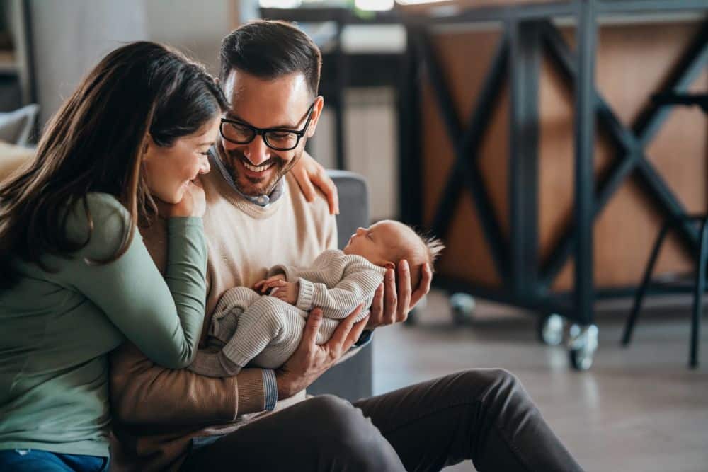 A happy man holding a newborn baby in his arms and looking at his wife leaning on his shoulder and looking at their baby