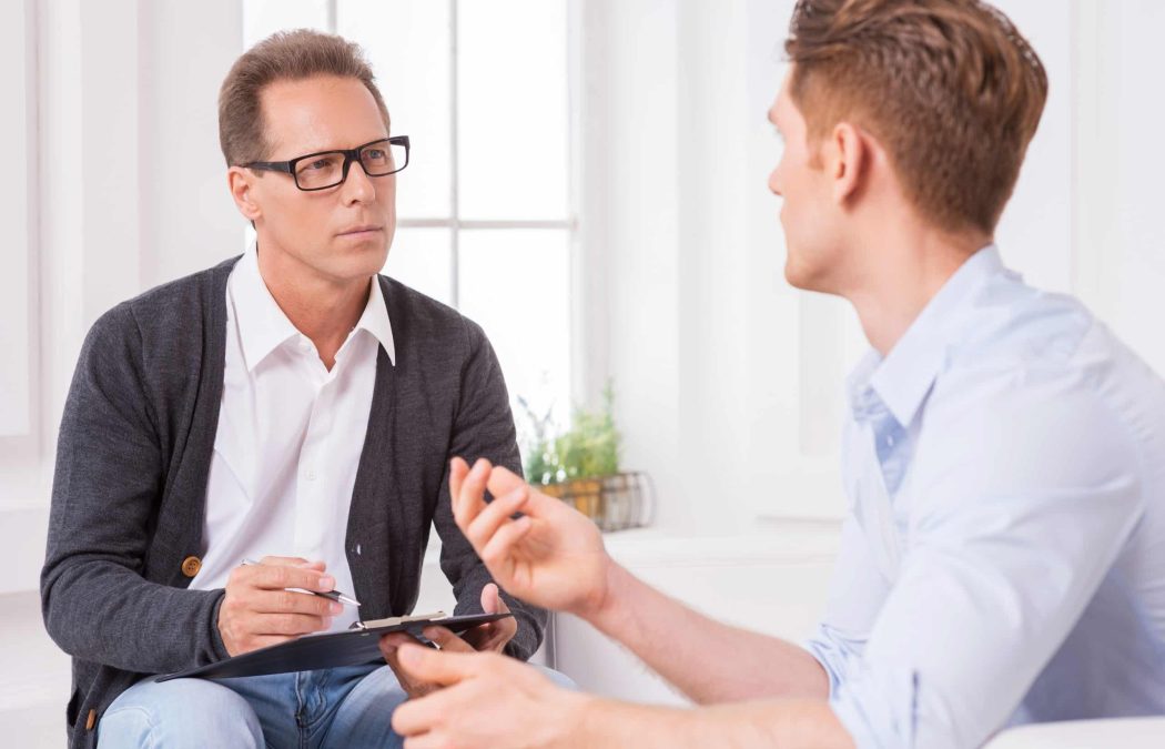 A therapist wearing glasses listens attentively and takes notes while a young man discusses concerns about adult circumcision during a counseling session in a bright office.