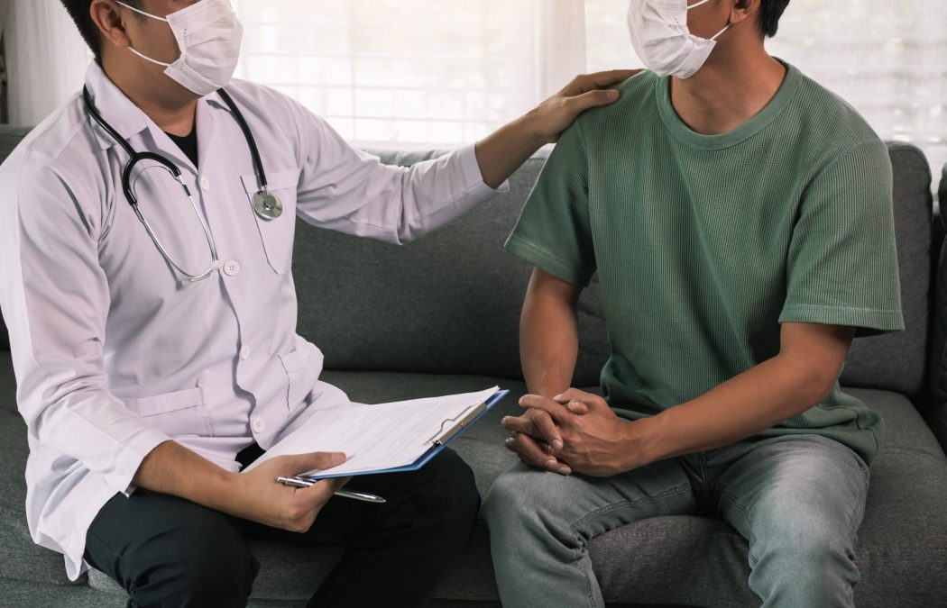 A doctor wearing a white coat and mask sits on a couch beside a masked patient