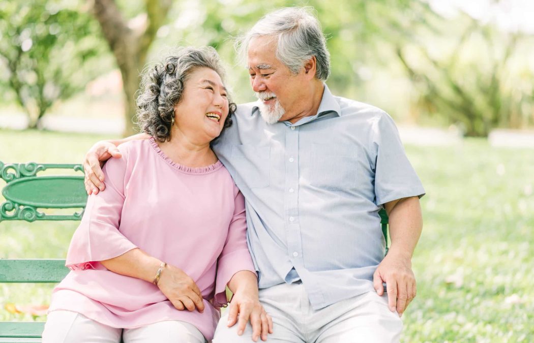 An older couple sits closely together on a green bench outdoors