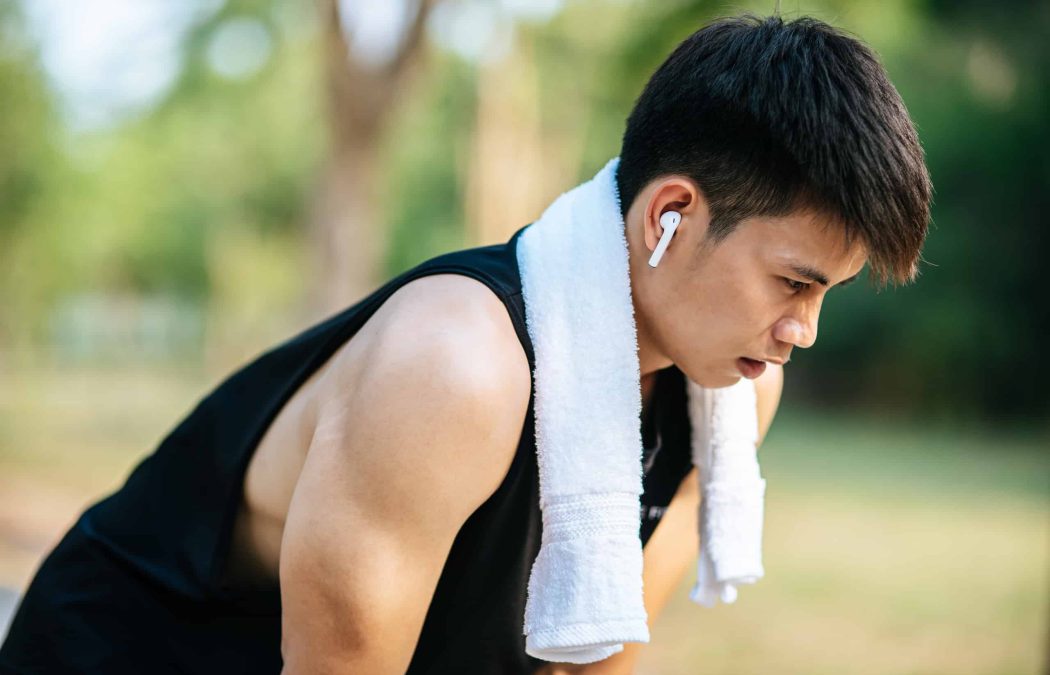 A young man wearing a sleeveless shirt and a white towel around his neck listens to wireless earbuds while leaning forward outdoors