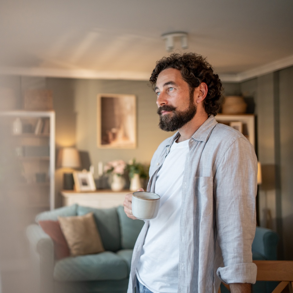 Thoughtful man with a beard, wearing a gray shirt and holding a coffee cup, gazes pensively out the window in his cozy living room, enjoying a quiet moment of reflection
