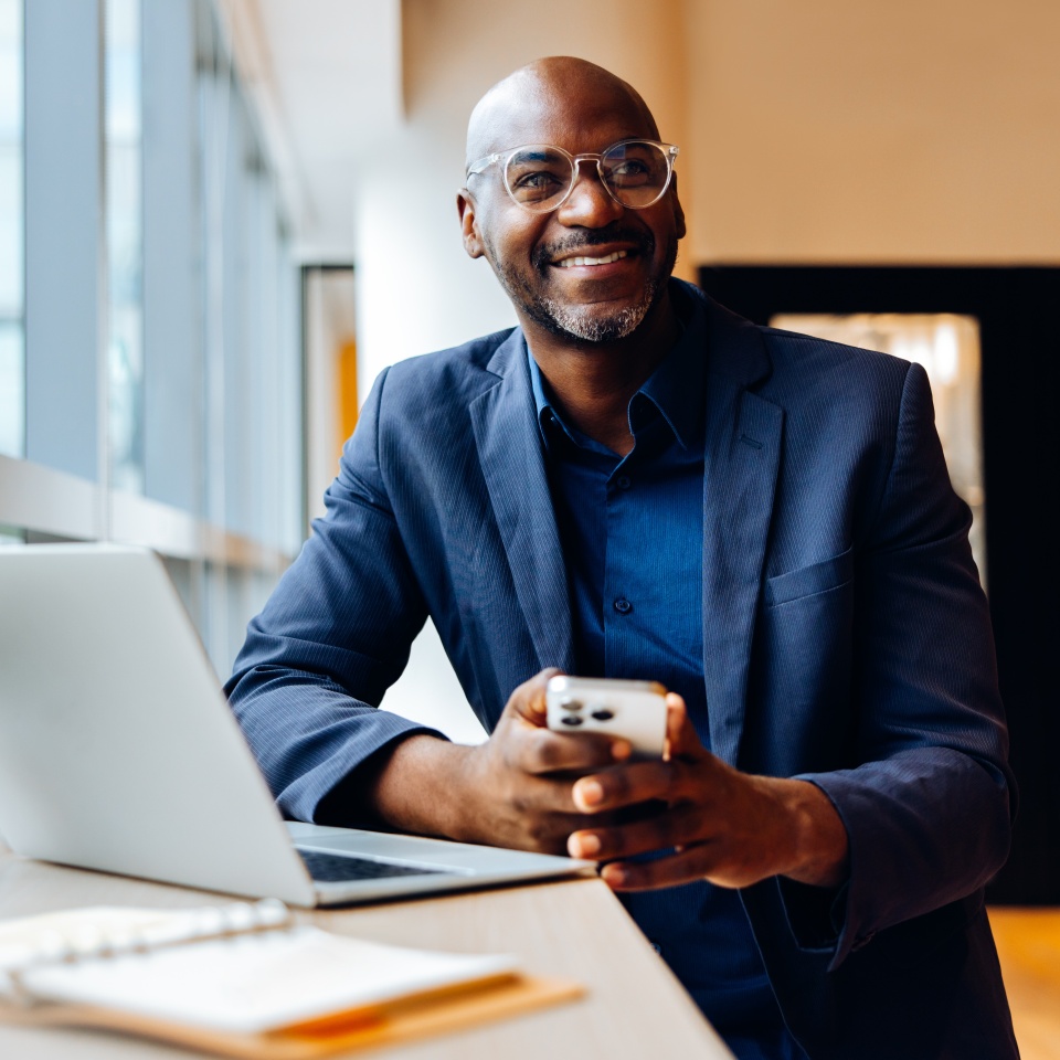 A cheerful man at his desk using a smartphone, with a laptop and papers, enjoying work.