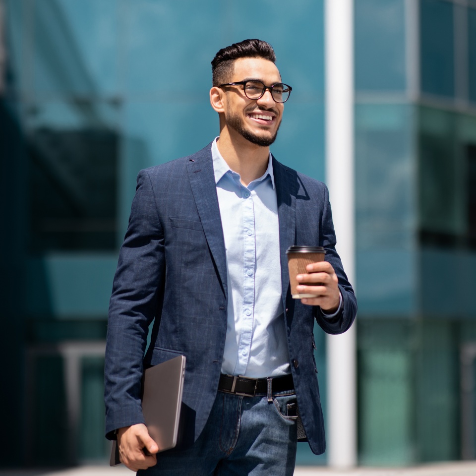 Cheerful middle eastern young businessman in glasses with laptop and coffee to go walking by street towards modern office building, going to job in the morning, panorama with copy space