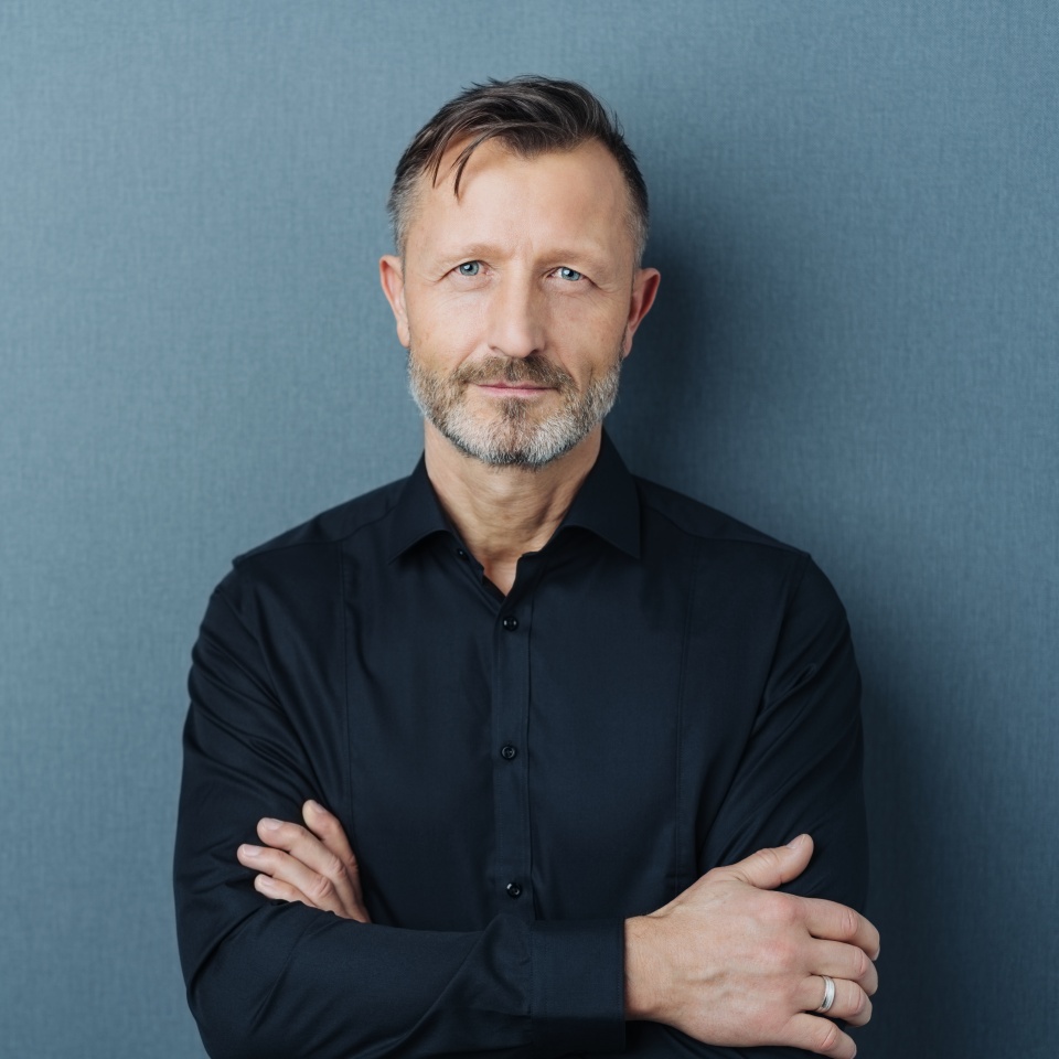 Serious middle-aged man with folded arms and a deadpan expression posing in front of a grey background with copy space