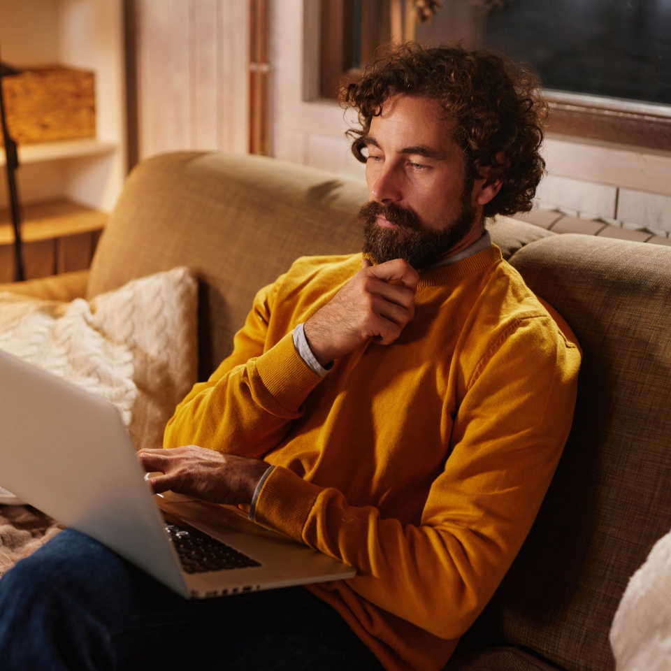 A man in a cozy sweater is sitting on a couch, focused on his laptop in a warm living room. Soft lighting creates a relaxed atmosphere as he ponders his work.