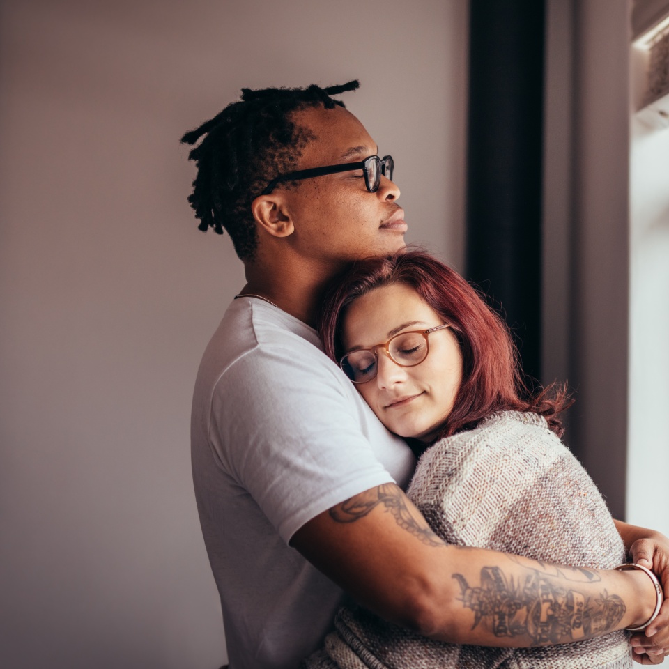 Interracial couple hugging each other while standing near window. African man embracing his caucasian girlfriend at home.