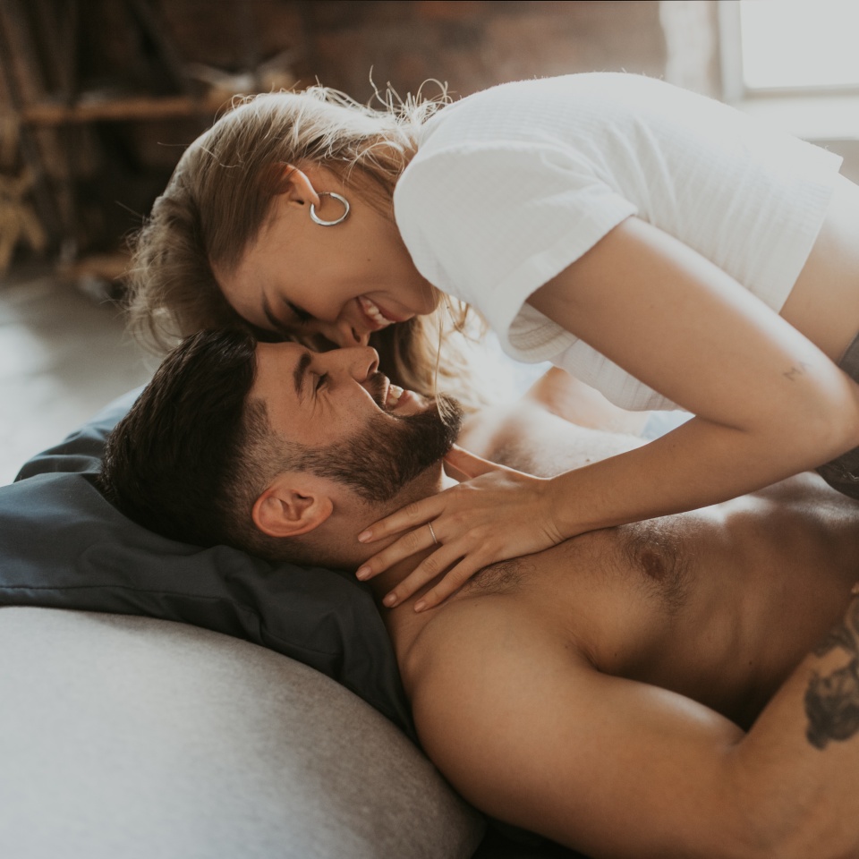 A woman leans over a shirtless man lying on a bed, both smiling and embracing in a cozy indoor setting.