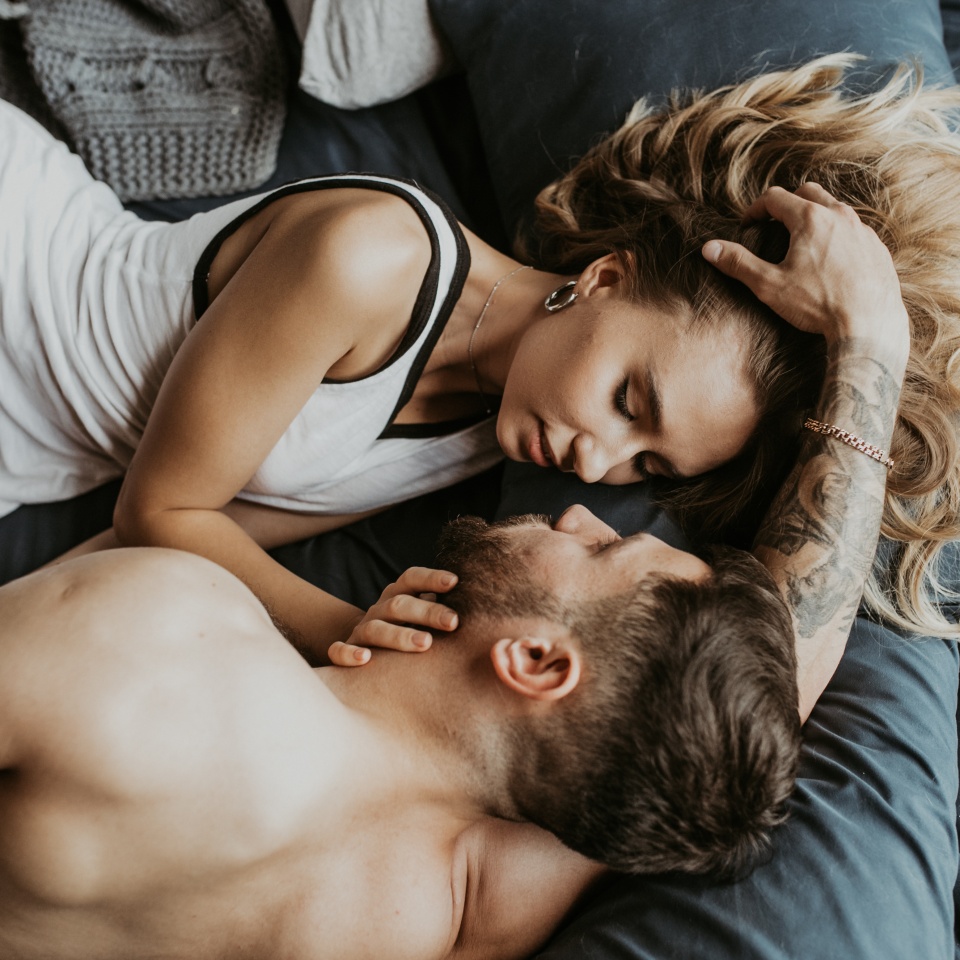A woman in a white tank top lies next to a shirtless man on a bed, touching his face and looking at him closely.
