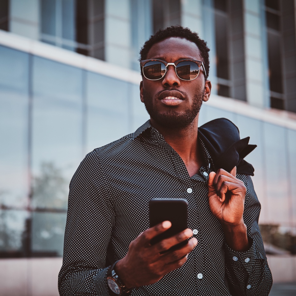 Portrait of pensive modern afro man with sunglasses and mobile phone over glass building background.