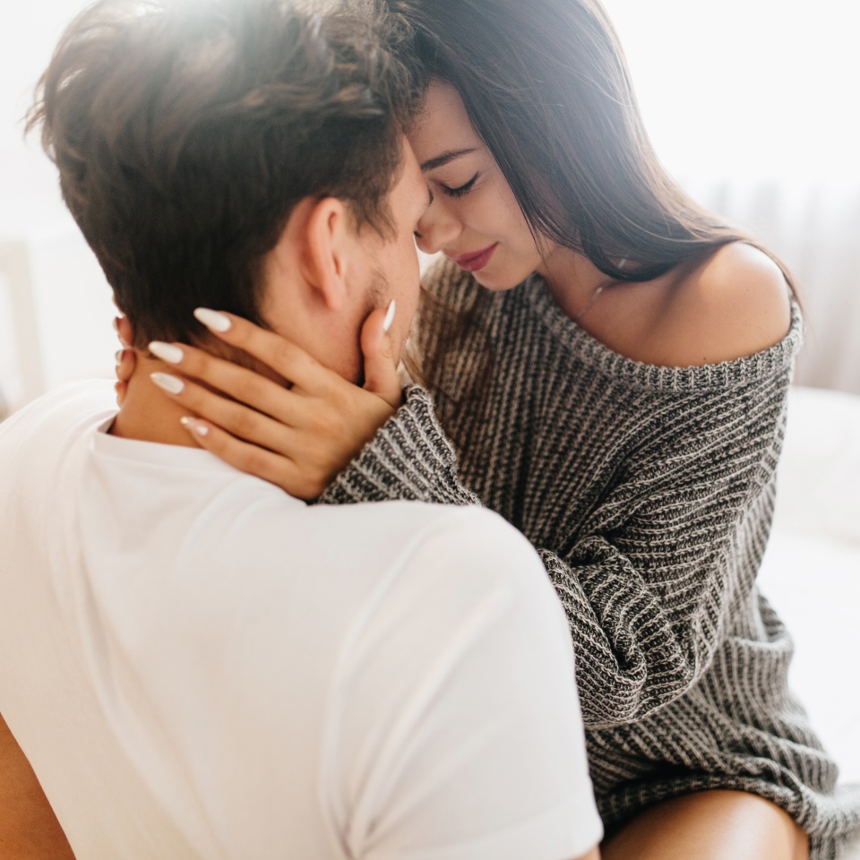 Glamorous tanned girl with white manicure resting with husband in sunday morning. Fascinating european woman holding boyfriend's face with tenderness, sitting on bed.