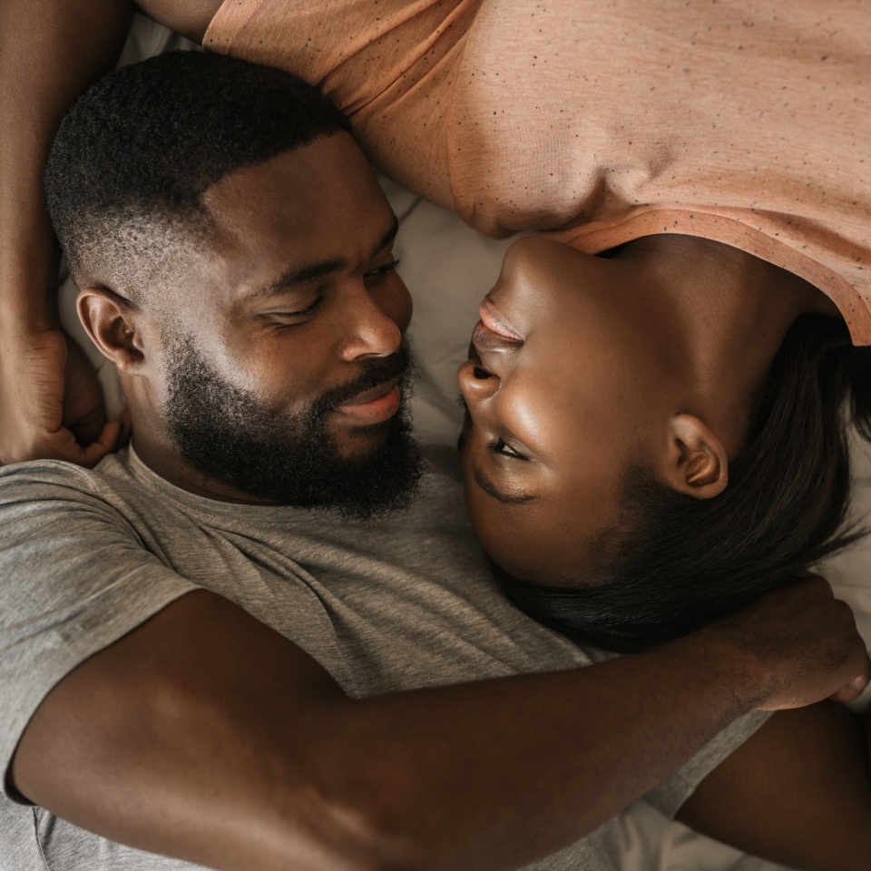 Affectionate young African American couple lovingly smiling at each other while lying face to face in bed together in the morning