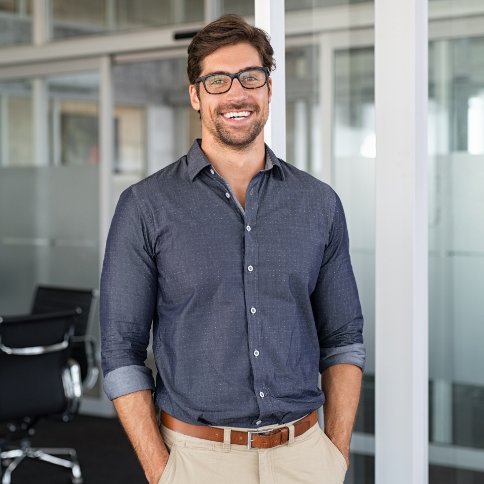 Portrait of young businessman wearing eyeglasses and standing outside conference room. Portrait of happy business man wearing spectacles and looking at camera with copy space. Satisfied proud man feeling confident in a modern office.