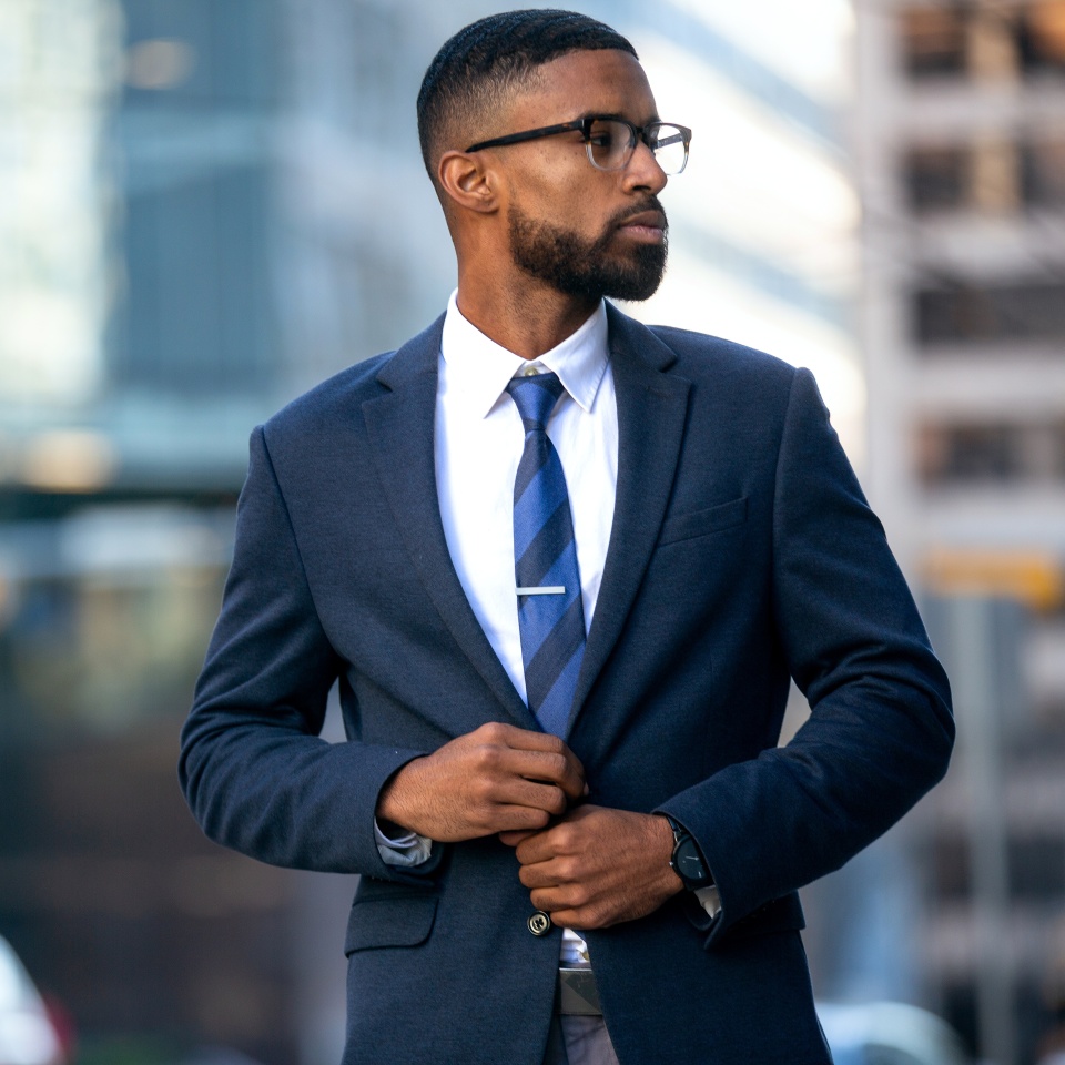 A man in a dark suit, white shirt, and striped tie stands outdoors in an urban setting, adjusting his jacket and looking to the side.