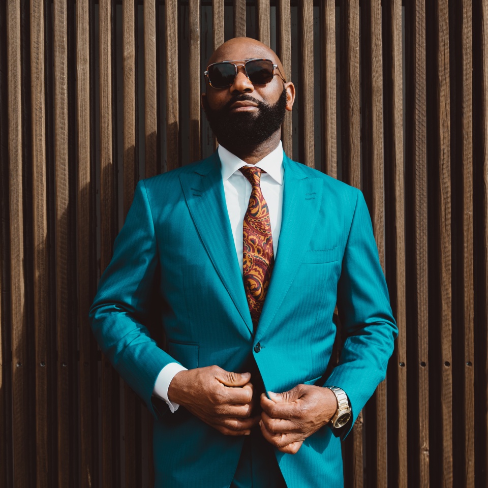 A handsome mature bald bearded African man in a sunglasses and a fashionable blue or teal costume with a tie is standing in front of a wall made of striped wooden timbers and fastening a suit button