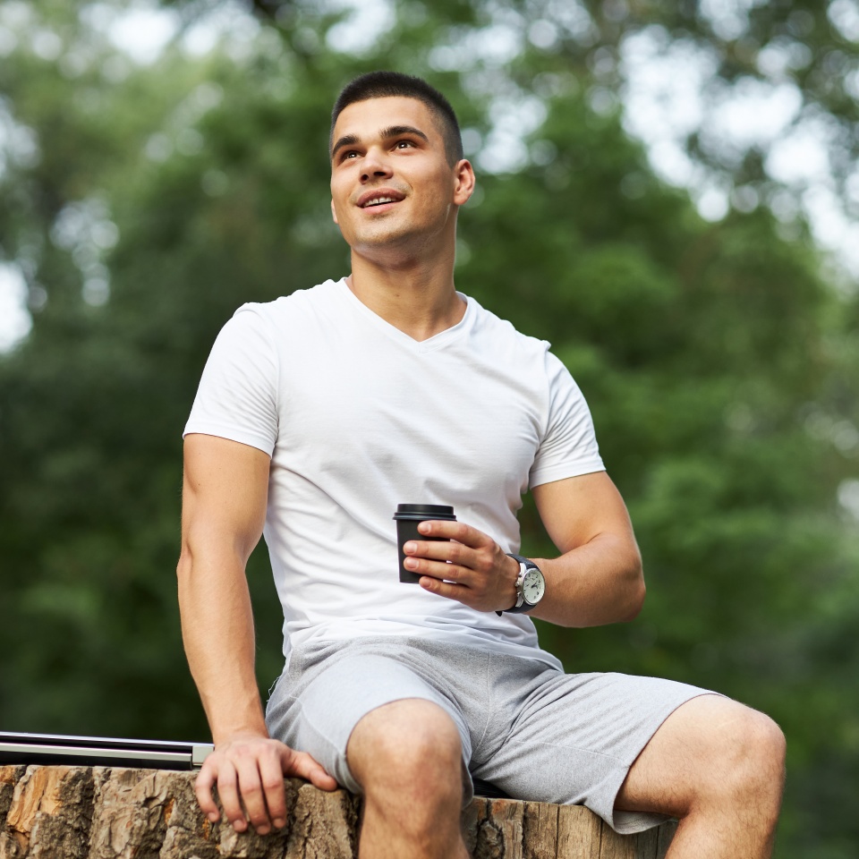 A young man sits in the park on a large stump with coffee in his hands. Shallow depth of field with emphasis on it.