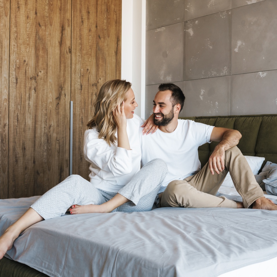 Image of young cheerful couple smiling and hugging while sitting on bed at home