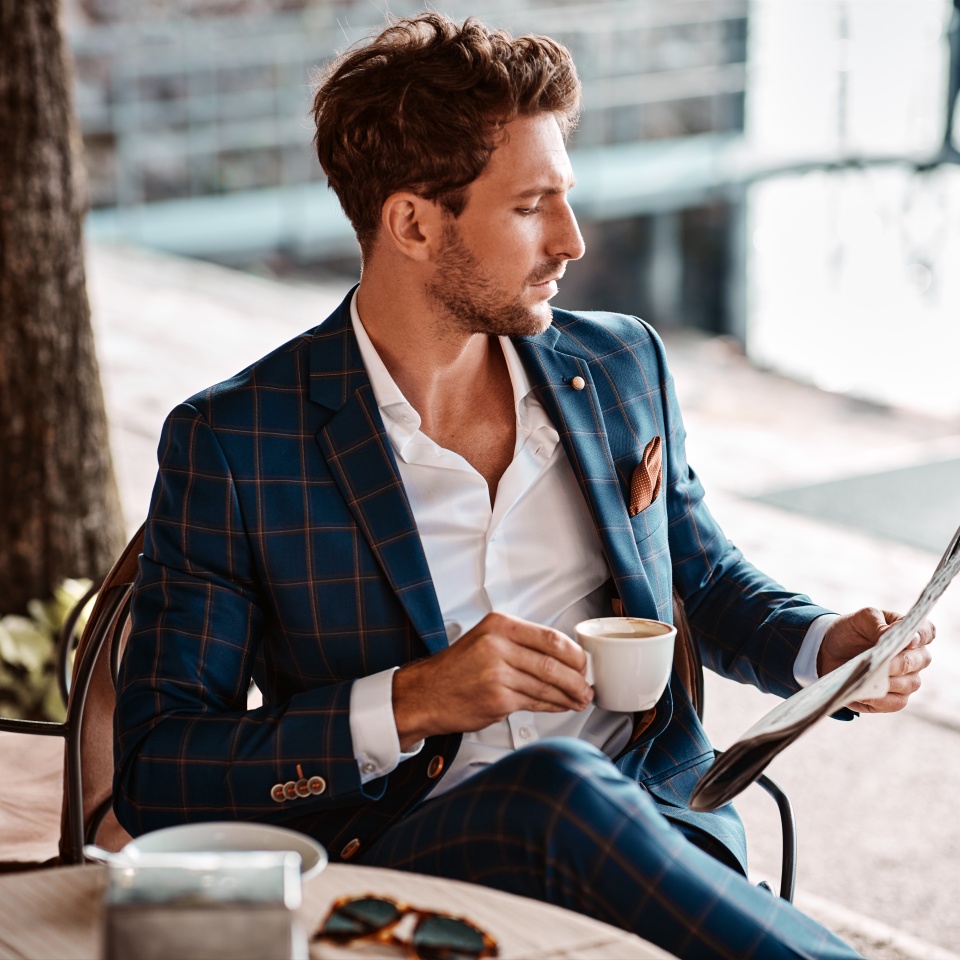 Man in a blue checkered suit sits outdoors, holding a cup and reading a newspaper at a café table.