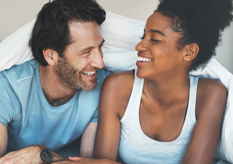 Happy, carefree and laughing couple having fun lying in bed together. Interracial husband and wife bonding and showing affection while talking. Smiling lovers enjoying time indoors being playful.