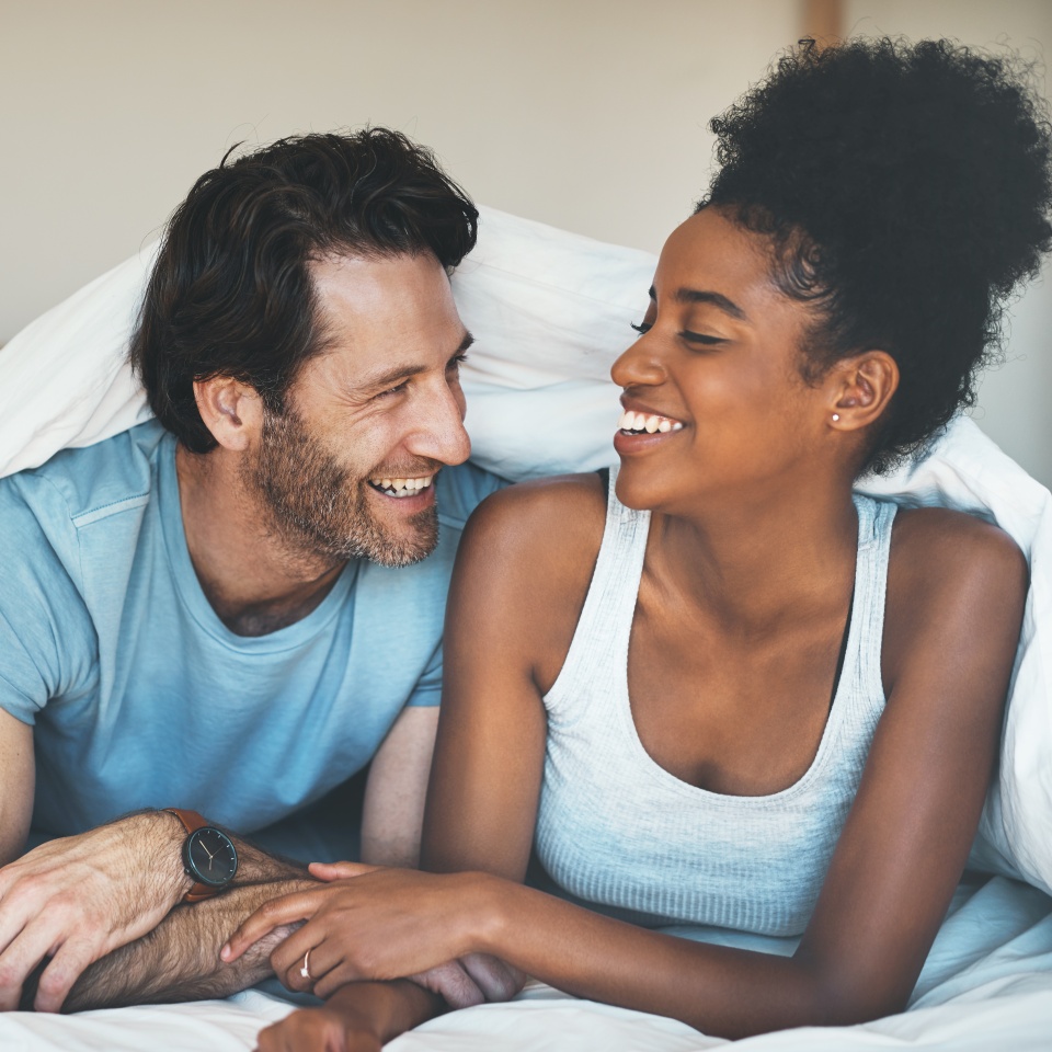 Happy, carefree and laughing couple having fun lying in bed together. Interracial husband and wife bonding and showing affection while talking. Smiling lovers enjoying time indoors being playful.