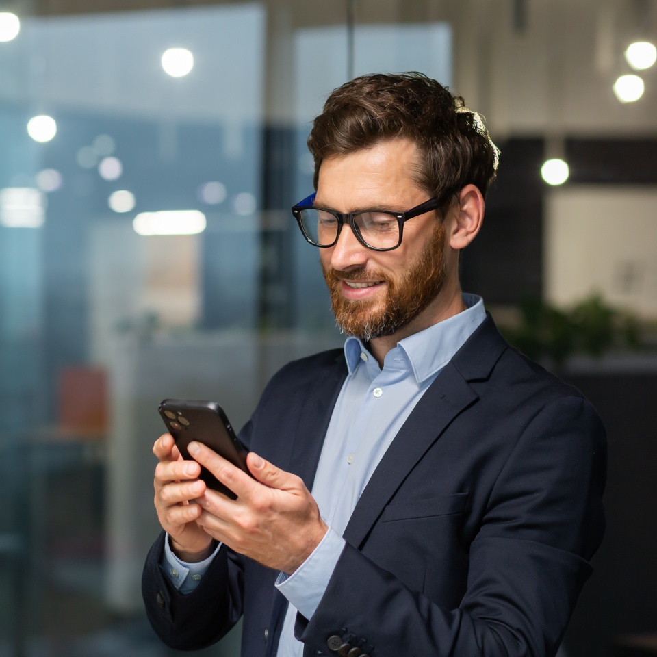 Successful financier investor works inside office at work, businessman in business suit uses telephone near window, man smiles and reads good news online from smartphone.