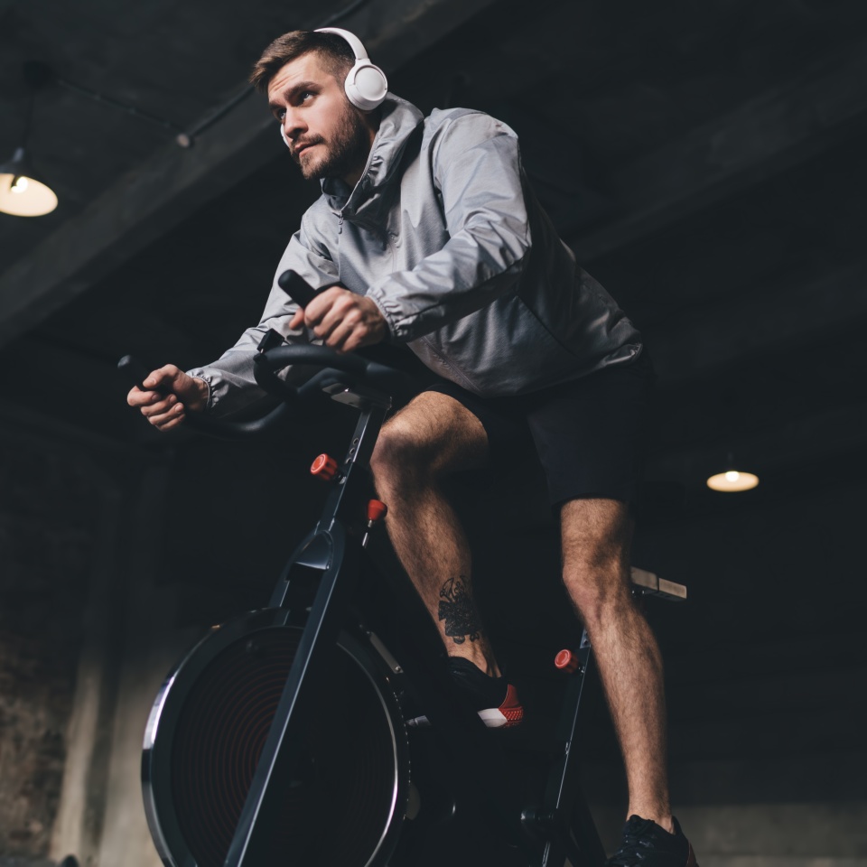 Low angle of young male in headphones looking away while sitting on spinning stationary cycle and listening to music in wireless headphones while doing cardio workout in gym