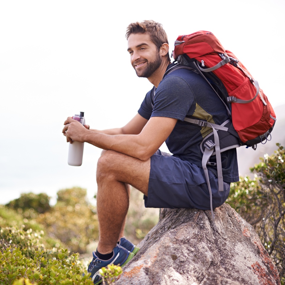 Hiking, mountain and man rest on a rock thinking after exercise, workout and fitness in nature for wellness. Travel, vacation and male person or athlete smile at a view after training and trekking.