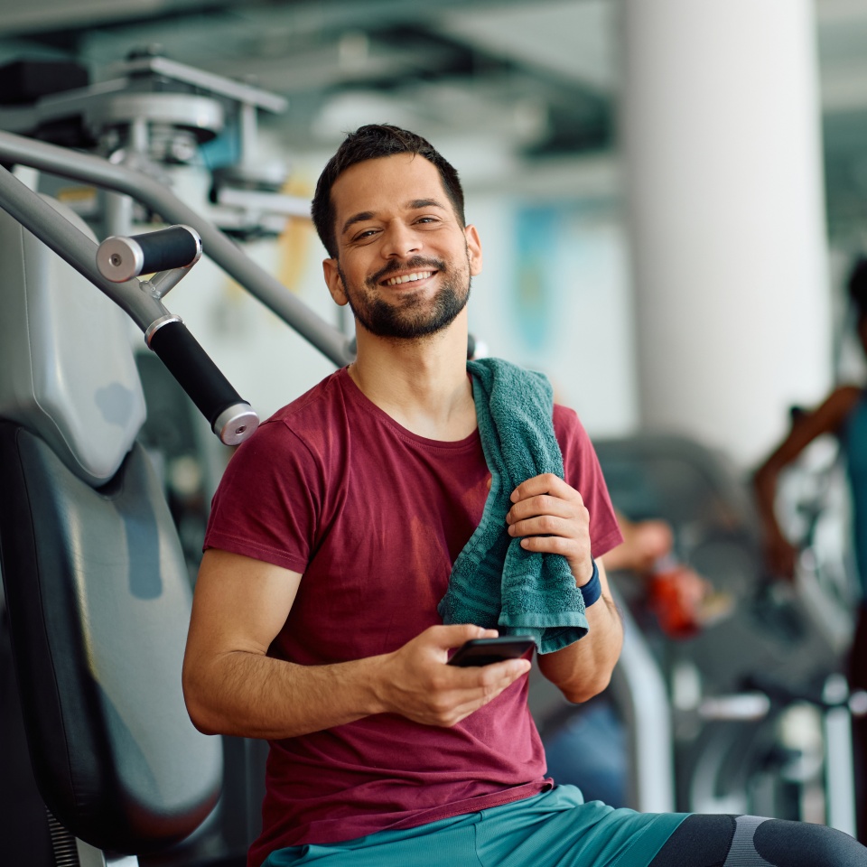 Man in gym clothes sits on exercise equipment, smiling and holding a smartphone and a teal towel, with gym equipment visible in the background.