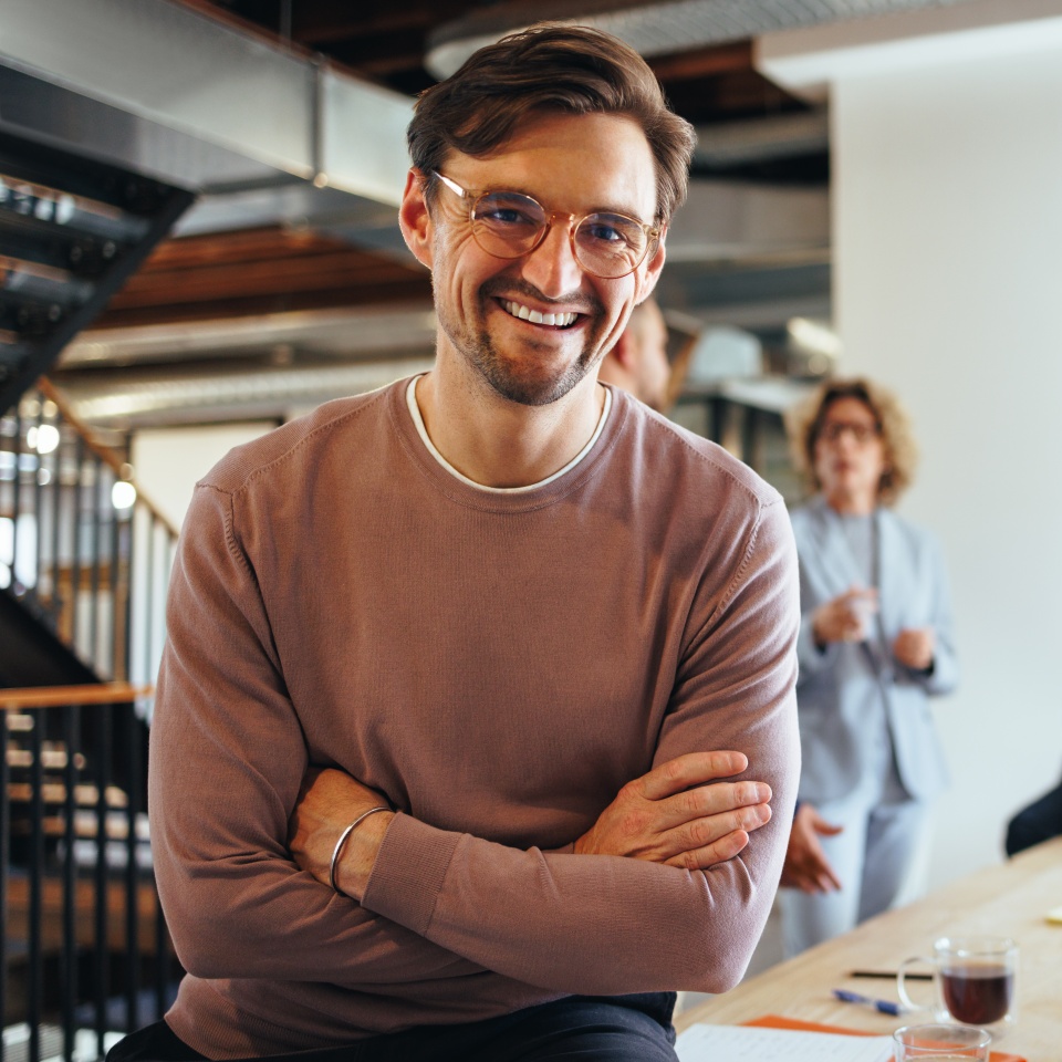 Successful business man sitting in a boardroom with his team in the background. Happy business professional looking at the camera in a meeting. Business people working on a project.