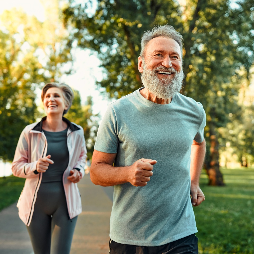 Running in park at morning time. Cheerful husband and wife competing together and jogging on fresh air. Active people wearing sport clothes doing cardio for good health and staying fit.