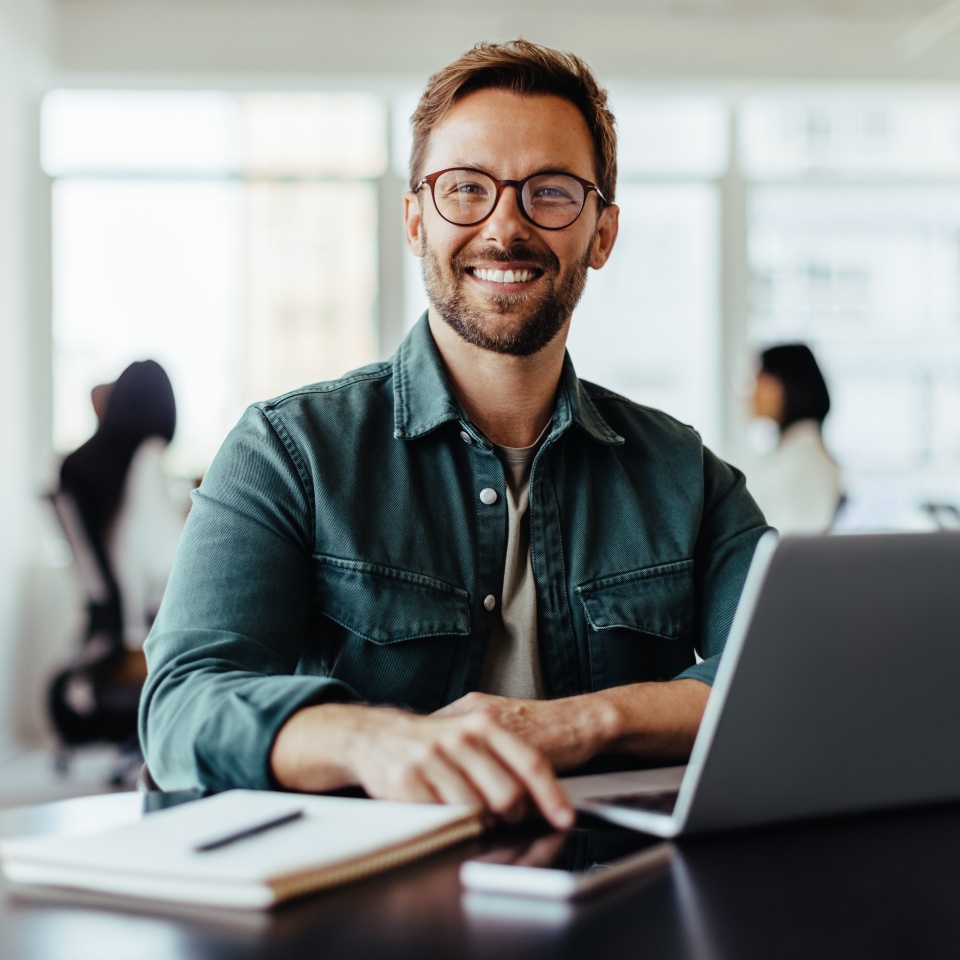 Portrait of a business man sitting in an office with his colleagues in the background. Happy business man working in a co-working office.