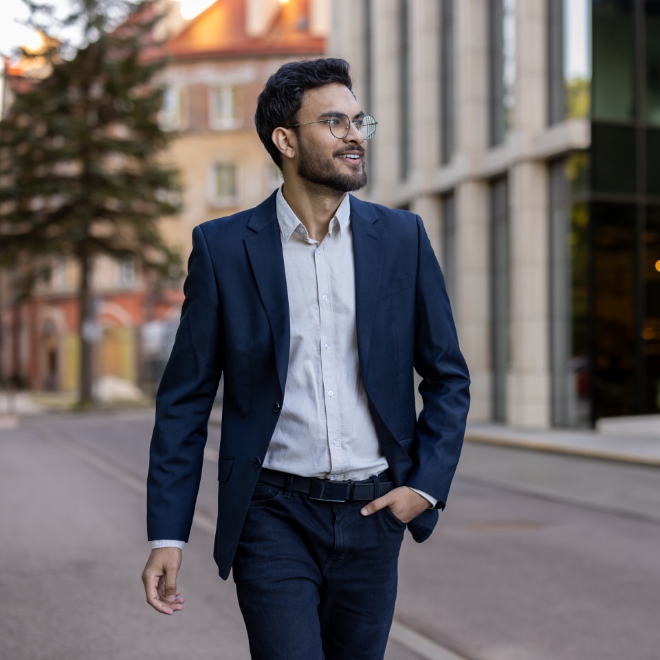 Confident smiling businessman wearing blazer and glasses walking in urban city street. Professional young man enjoying his day. Concept of success, confidence, business attire.