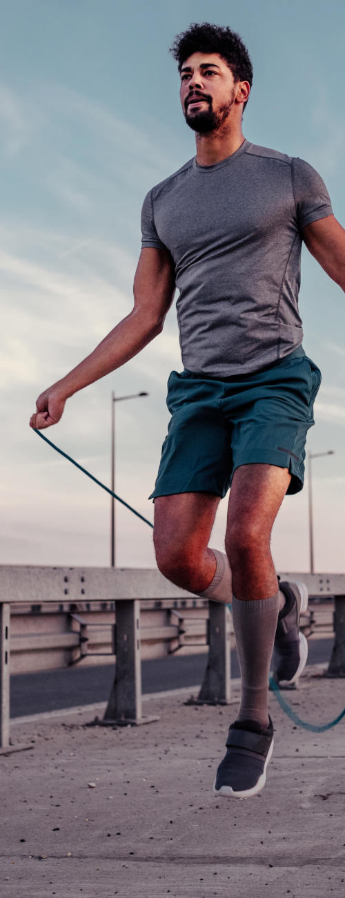 A man in athletic wear jumps rope outdoors on a concrete surface near a metal railing, with a cloudy sky in the background.